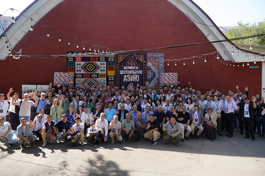 A large group of church leaders gathered outdoors in front of a red building in Almaty, Kazakhstan.